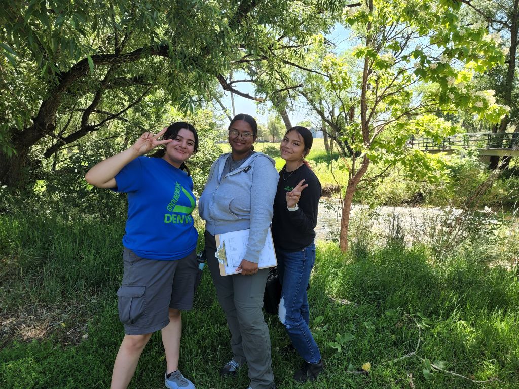 Photo of three young people in Groundwork Denver tee shirts smiling and making peace signs as they stand under a tree at the bank of a river on a sunny day.