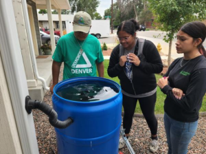 Three youth in Groundwork Denver apparel surround a rain barrel they installed. They are looking at the water, which is filled to the brim.