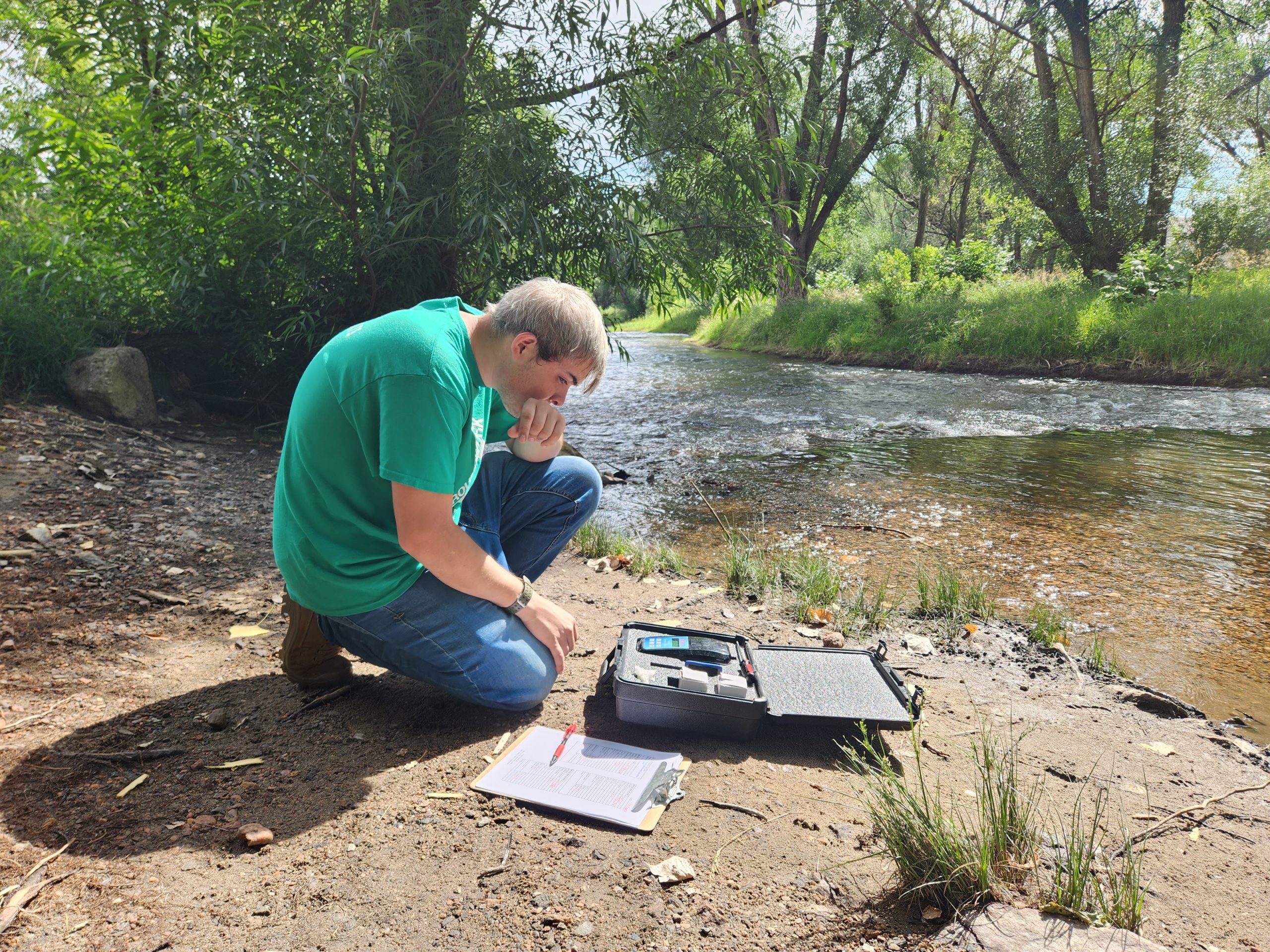 A young person in a Groundwork Denver tee sits on the bank of Bear Creek taking measurement of contamination levels.