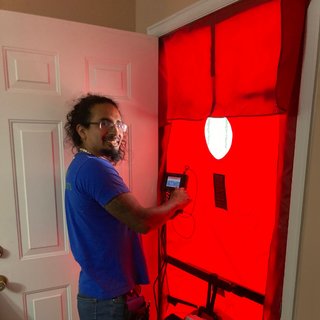 Energy auditor takes a reading from a blower door test. He's smiling and holding a monitoring device in front of a door with a large red panel that seals the home for the test.