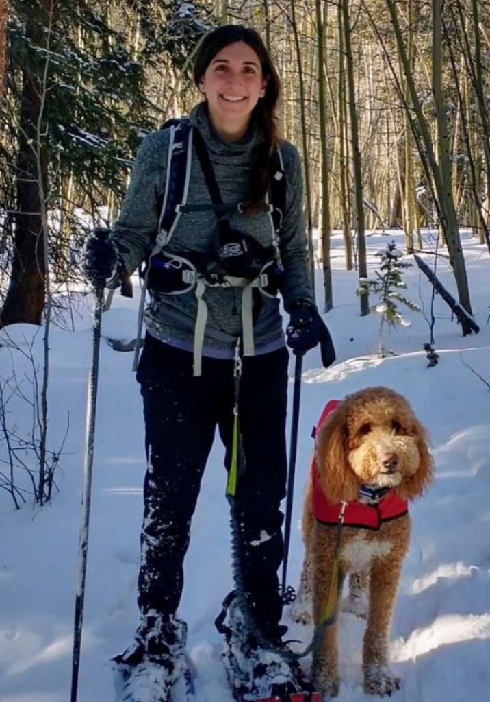 Elissa poses with her dog in snowy woods. She's smiling and wearing snowshoes.